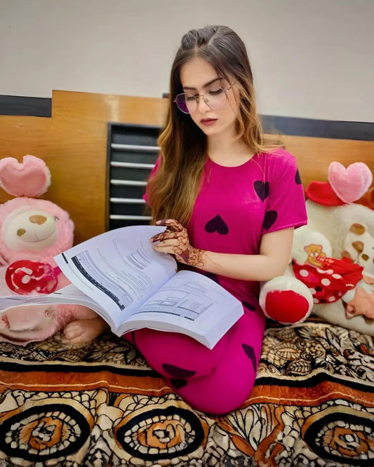 Woman in pink shirt with heart patterns reading a book on a bed with stuffed animals.